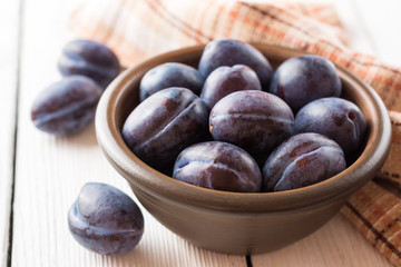 Fresh plums in ceramic bowl on white wooden background.