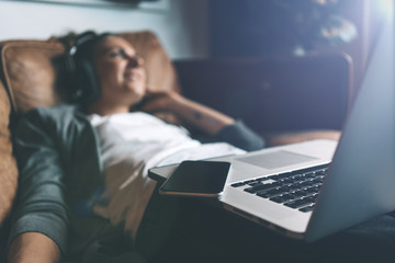 Attractive young happy woman listening to music in headphones with pleasure at coworking office, lying on sofa, using laptop and mobile phone and resting.