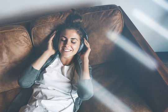 Attractive young happy girl listening to music from headphones with pleasure at coworking office, lying on sofa and resting.