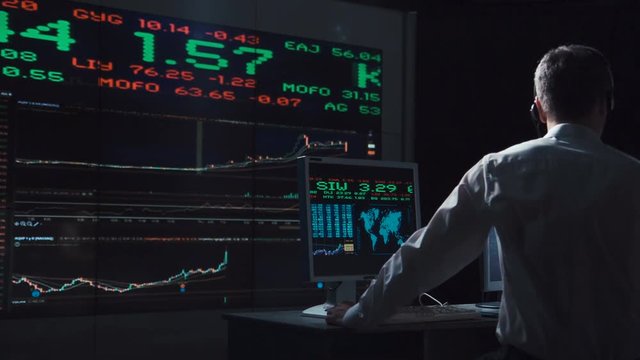 Adult man working in office of exchange and driving lively trade while speaking with headset.