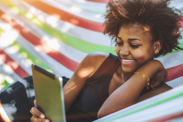 Cute cheerful teenage Brazilian female with African hair is laying in colorful striped hammock located in public park on warm sunny day and smiling while reading funny memes on her digital tablet - Powered by Adobe