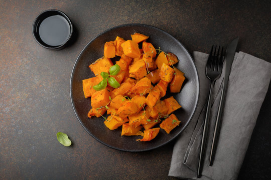 Fried pumpkin slices with herbs and pepper, olive oil in a black ceramic plate on  dark concrete or stone background. Selective focus. Top view.