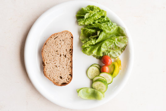 High Angle View Of White Plate With Brown Bread And Healthy Salad Including Lettuce, Tomato And Cucumber On Limestone Background