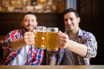 Having a pint with friend. Two cheerful young men toasting with beer while sitting together at the bar
