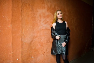 Blonde fashionable girl in long black leather coat posed near street tonel with orange wall.