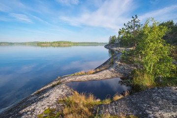 Fototapeta na wymiar Landscape on the beautiful stone shore of the lake with islands in the summer morning with a beautiful sky and reflections. Ladoga Lake, Karelia, the island of Koyonsaari