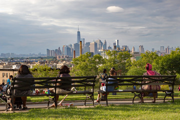New York's diversity represented by people sat on a bench in Sunset Park, Brooklyn, admiring the view of Lower Manhattan on a sunny summer's day - obrazy, fototapety, plakaty