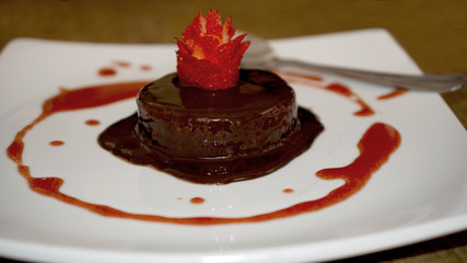 Chocolate fudge covered with chocolate sauce and decorated with sculptured strawberry and spoon on white plate on wooden background