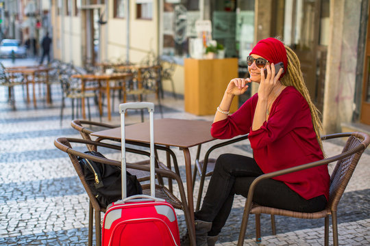 A young woman with a red suitcase sitting in a street cafe talking on the phone.