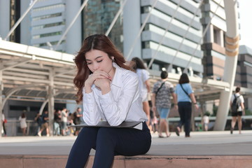 Portrait of pretty young Asian business woman thinking and sitting on stairway in urban building background.