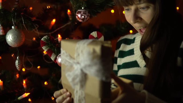 A young woman opens a gift box, the brunette sits under a Christmas tree on a background of yellow lights in the early morning or at night indoors.