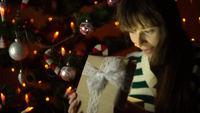 A young woman opens a gift box, the brunette sits under a Christmas tree on a background of yellow lights in the early morning or at night indoors, close-up.