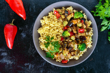 Light healthy dietary vegan dish: couscous and vegetables (zucchini, eggplant, carrots, sweet peppers, onions, parsley) on a black background. The top view.