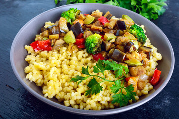 Light healthy dietary vegan dish: couscous and vegetables (zucchini, eggplant, carrots, sweet peppers, onions, parsley) on a black background.