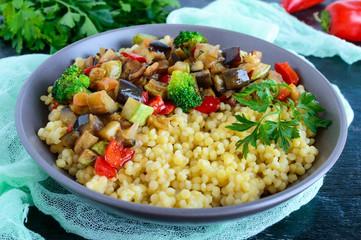 Light healthy dietary vegan dish: couscous and vegetables (zucchini, eggplant, carrots, sweet peppers, onions, parsley) on a black background.