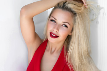 Fototapeta premium Fashion image of young smiling beautiful woman in dress.Close-up portrait of young elegant blonde woman in red dress and red lips touching her long blonde hair. Fashion indoor shot