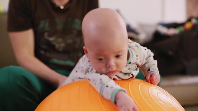 The father rolls a newborn on a fitness ball, is engaged in sports and massage. Cute baby lying on the orange fitball on the blue background. Concept of caring for the baby's health.