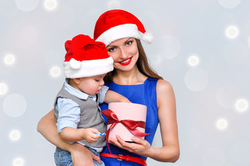 Mother holding her child and shows him a box with a red bow. A little boy takes out the gift from the box. Both in Santa hats