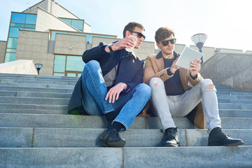 Portrait of two handsome young men using digital tablet outdoors sitting on steps of modern glass building and enjoying coffee