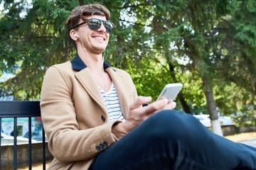 Portrait of happy young man smiling while using smartphone and listening to music outdoors sitting on park bench enjoying sunny autumn day