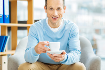 Positive emotions. Happy delighted joyful man holding a cup of coffee and smiling while being in a positive mood
