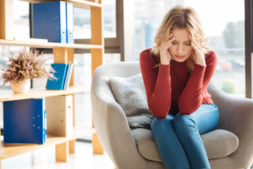 Obraz premium Strong headache. Unhappy cheerless depressed woman sitting in the armchair and holding her temples while having a strong headache