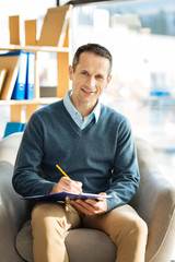 Professional psychologist. Cheerful happy nice man sitting in the armchair and taking notes while working as a psychologist