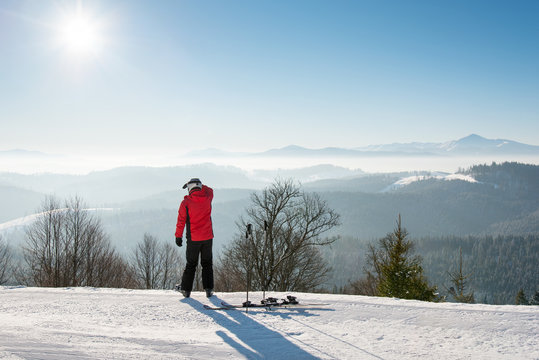 Male skier resting after the ride standing with his back to the camera on top of the ski slope looking around enjoying view of snowy mountains on a sunny winter day seasonal recreational active
