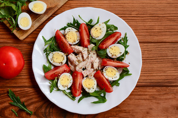 Easy vegetable chicken salad idea for lunch or dinner. Home salad with fresh tomatoes, arugula, quail eggs, chicken breast and spices on a white plate and a wooden table. Closeup. Top view