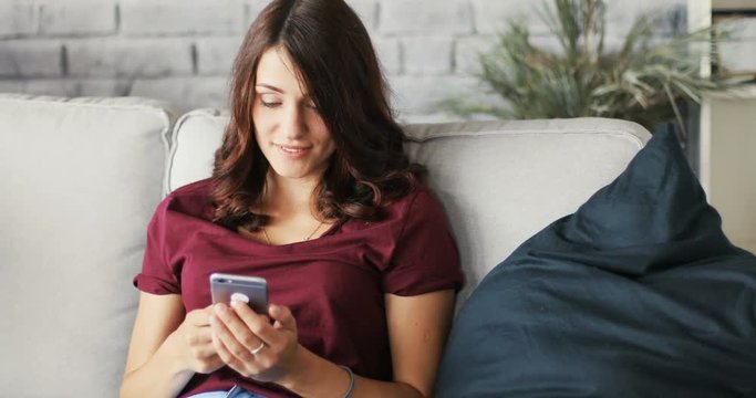 Portrait of a young attractive woman using the phone sitting on the couch in home smiling message smartphone apartment, cellphone communication internet sms typing Slow Motion Shot on RED EPIC camera