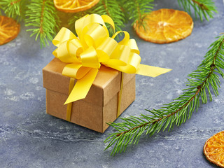 Fototapeta na wymiar Christmas Kraft gift box with yellow bow on a grey stone background. Green spruce branches and decoration of dried oranges.