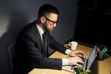 Attentive bearded man in glasses working on project late at night, typing messages, writhing emails using laptop computer. Forgotten cold cup of coffee or tea at his hand.