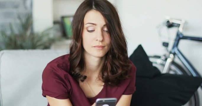 Close up of a young attractive woman using the phone sitting on the couch in home smiling message smartphone apartment, cellphone communication internet sms typing Slow Motion Shot on RED EPIC camera
