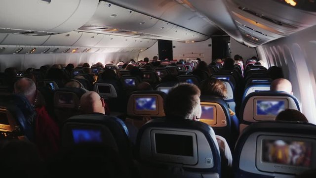 In the cabin of a large airliner. Passengers in comfortable armchairs watching entertaining programs on screens