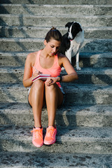 Fitness woman taking workout routine notes and timing training. Female athlete sitting on stairs with her dog.