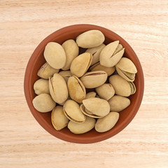 Top view of a small red clay bowl filled with roasted pistachio nuts on a brown wood table.
