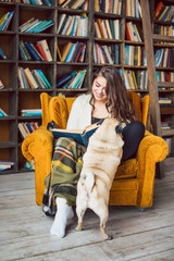 Beautiful young woman reading in home library and her funny cute dog pug asking playing