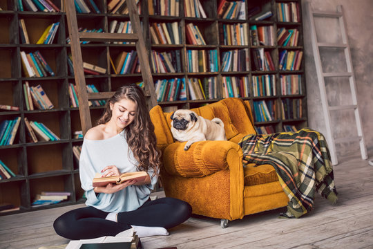 Beautiful reading woman in library and her funny pug dog. Happy relaxed home weekend. Cozy home atmosphere. 