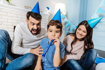 Party mood. Handsome little boy blowing into a party horn while his mother and father cheering him and laughing