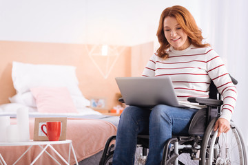 Fototapeta na wymiar Being a freelancer. Beautiful alert blond woman of middle age smiling and holding laptop while sitting in the wheelchair