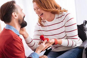 Over the moon. Happy smiling bearded man making a proposal to an attractive alert blond handicapped woman laughing and caressing him and holding his hand with the ring while sitting in a wheelchair