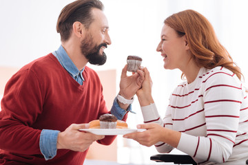 Having lunch. Happy bearded man and blond woman smiling and looking at each other while holding together a plate with some cookies