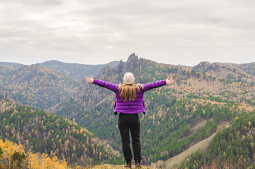 Obraz premium A girl in a lilac jacket stretching her arms on a mountain, a view of the mountains and an autumn forest by a cloudy day, free space for text