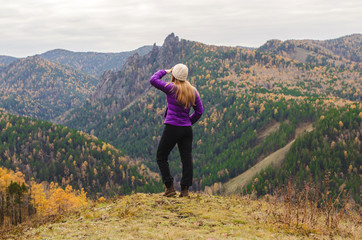 Fototapeta premium A girl in a lilac jacket looks out into the distance on a mountain, a view of the mountains and an autumnal forest by an overcast day. Free space for text