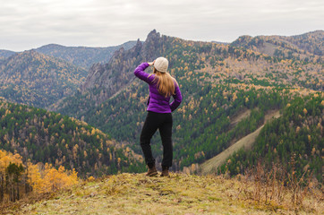 A girl in a lilac jacket looks out into the distance on a mountain, a view of the mountains and an autumnal forest by an overcast day. Free space for text