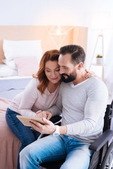 Obraz premium We love hugging. Cheerful smiling blond woman and a bearded disabled man holding hands and looking at the picture while the man sitting in the wheelchair and the woman sitting on the couch