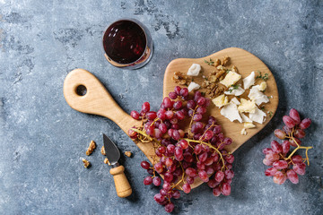 Wooden serving board with fresh red grapes, walnuts, goat and cheddar cheese, with cheese knife and glass of red wine over blue texture background. Top view, copy space.