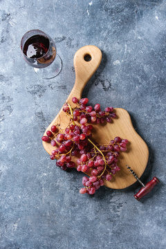 Wooden serving board with fresh red grapes, corkscrew and glass of red wine over blue texture background. Top view, copy space.