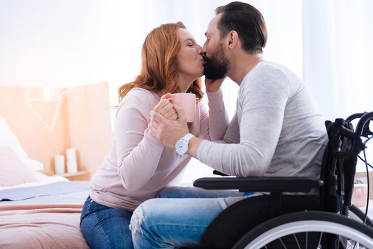 We love kissing. Blond woman of middle age and a bearded disabled man kissing while the woman holding a cup and the man sitting in a wheelchair
