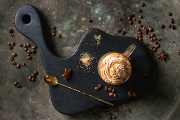 Glass of spicy pumpkin latte with whipped cream and cinnamon standing on black serving board. Coffee beans and spices above. Dark background. Top view, space.